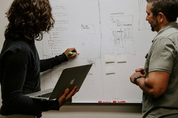 Photo by Kaleidico on Unsplash. Photo of two men with one holding a laptop and writing a process on a whiteboard